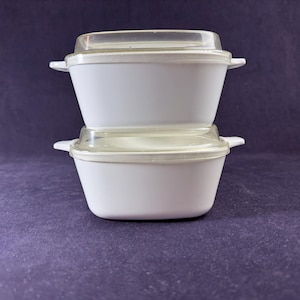 May include: Two white, rectangular casserole dishes with clear lids are stacked on top of each other. Each dish has small handles on either side. The background is a solid, dark purple color.