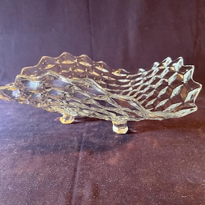 May include: Clear glass serving dish with a textured, diamond-patterned design. The dish has a scalloped edge and rests on three small feet. The overall shape is elongated and reminiscent of a seashell.