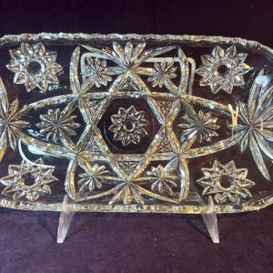 May include: A clear, rectangular glass serving tray with a starburst and geometric design. The tray is supported by a clear stand, showcasing the intricate patterns and textures of the cut glass. The tray's edges are scalloped.