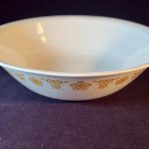 May include: White ceramic bowl with a gold floral pattern around the rim. The bowl has a wide, shallow shape and a smooth, glossy finish. The gold design features stylized flowers and other decorative elements.