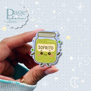 May include: A colorful sticker featuring a cartoon jar of sofrito. The jar is green with a label that reads "SOFRITO" and has a smiling face. The sticker is held by a hand against a light blue background with stars and moons.