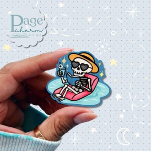 May include: A die-cut sticker featuring a skeleton wearing a yellow sun hat and heart-shaped sunglasses, relaxing on a pink pool float in blue water while reading a blue book and holding a drink with an eyeball garnish.