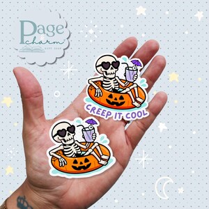 May include: Two stickers featuring skeleton characters wearing heart-shaped sunglasses, relaxing in orange pumpkin-shaped floats, and holding drinks with umbrellas. The text "Creep It Cool" is written on the stickers. The stickers are on a hand.