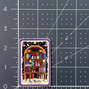 May include: A rectangular sticker featuring a bookshelf illustration with the text The Reader. The sticker is placed on a dark gray cutting mat with white grid lines, showing it measures approximately 2.5 inches in height.