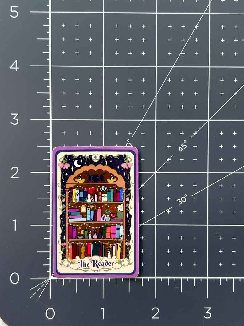 May include: A rectangular sticker featuring a bookshelf illustration with the text The Reader. The sticker is placed on a dark gray cutting mat with white grid lines, showing it measures approximately 2.5 inches in height.