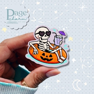 May include: A sticker featuring a cartoon skeleton wearing heart-shaped sunglasses, relaxing in an orange pumpkin-shaped float. The skeleton holds a purple drink with an eyeball and a tiny purple umbrella. The sticker has the text "Page & Charm Gift Shop".