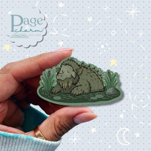 May include: A die-cut sticker featuring a green, shaggy, swamp-dwelling creature resting in dark water surrounded by cattails and lily pads. The illustration has a soft, hand-drawn aesthetic with muted earth tones and a dark green outline.