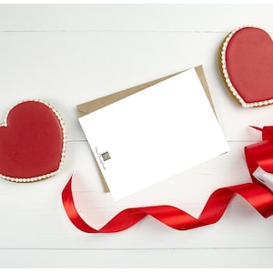 May include: A white card with a brown envelope, a red ribbon, a white gift box with a red ribbon, and two red heart-shaped cookies on a white wooden surface.