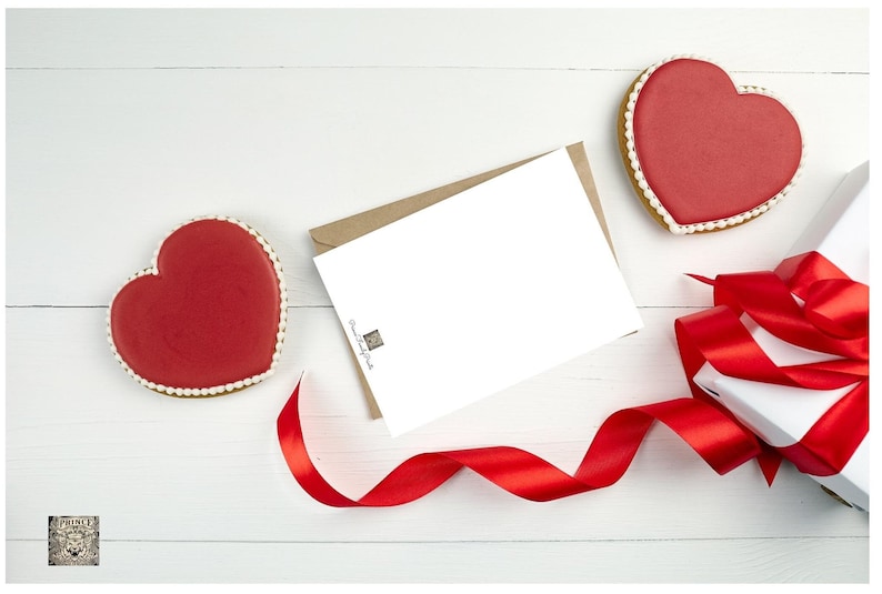 May include: A white card with a brown envelope, a red ribbon, a white gift box with a red ribbon, and two red heart-shaped cookies on a white wooden surface.