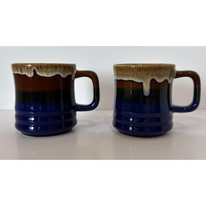 Puede incluir: Dos tazas de cerámica con un diseño horizontal de bloques de color y acabado brillante. Las tazas muestran bandas de color azul oscuro, negro y marrón, rematadas con un efecto de esmalte de goteo color crema en el borde y asa cuadrada.