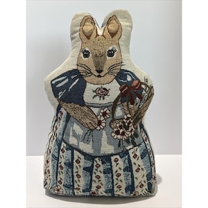 May include: A shaped decorative pillow featuring a woven tapestry design of a rabbit wearing a blue dress and apron. The rabbit holds a woven basket filled with white flowers. The fabric displays a detailed pattern of small red floral accents.