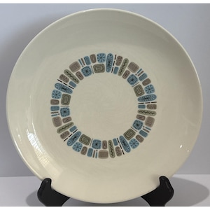 May include: A round, off-white ceramic dinner plate featuring a central circular pattern composed of small, repeating geometric shapes in shades of light blue, tan, and dark brown. The design includes stylized starbursts, rectangles, and dashes.