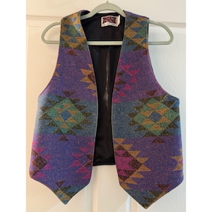 May include: A sleeveless vest featuring a vibrant purple base with a repeating geometric diamond pattern in shades of teal, magenta, and gold. The garment has a black interior lining and a white label at the neckline with the text "NEW SPIRIT CLOTHING".