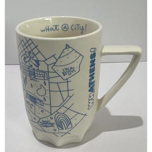 May include: A cream-colored ceramic mug featuring blue line-art illustrations of Athens landmarks, including the Kallimarmaro Stadium and Lycabettus. The text "WHAT ATHENS!" is printed vertically on the side, and "what A city!" is inside the rim.