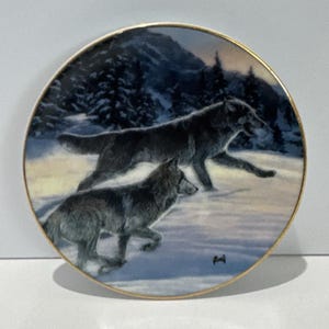 May include: A round decorative ceramic plate with a gold-toned rim featuring a detailed illustration of two grey wolves running through a snowy landscape. The background depicts dark evergreen trees and a mountain range under a twilight sky.