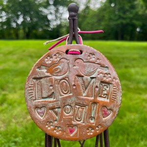 Puede incluir: Un adorno redondo de cerámica marrón con las palabras "LOVE YOU!" en letras en relieve. El adorno está decorado con dos pájaros, corazones y flores.