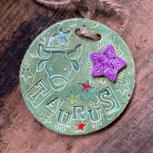 May include: A green ceramic ornament with the word "TAURUS" in a stylized font. The ornament features a bull, stars, and a purple star with a five-pointed star inside.