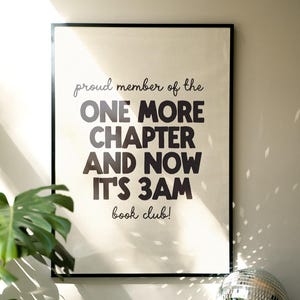 May include: A framed print with the text "proud member of the ONE MORE CHAPTER AND NOW IT'S 3AM book club!" is displayed. The print is in a black frame and is on a wall. A disco ball and a plant are on a wooden surface below the print.