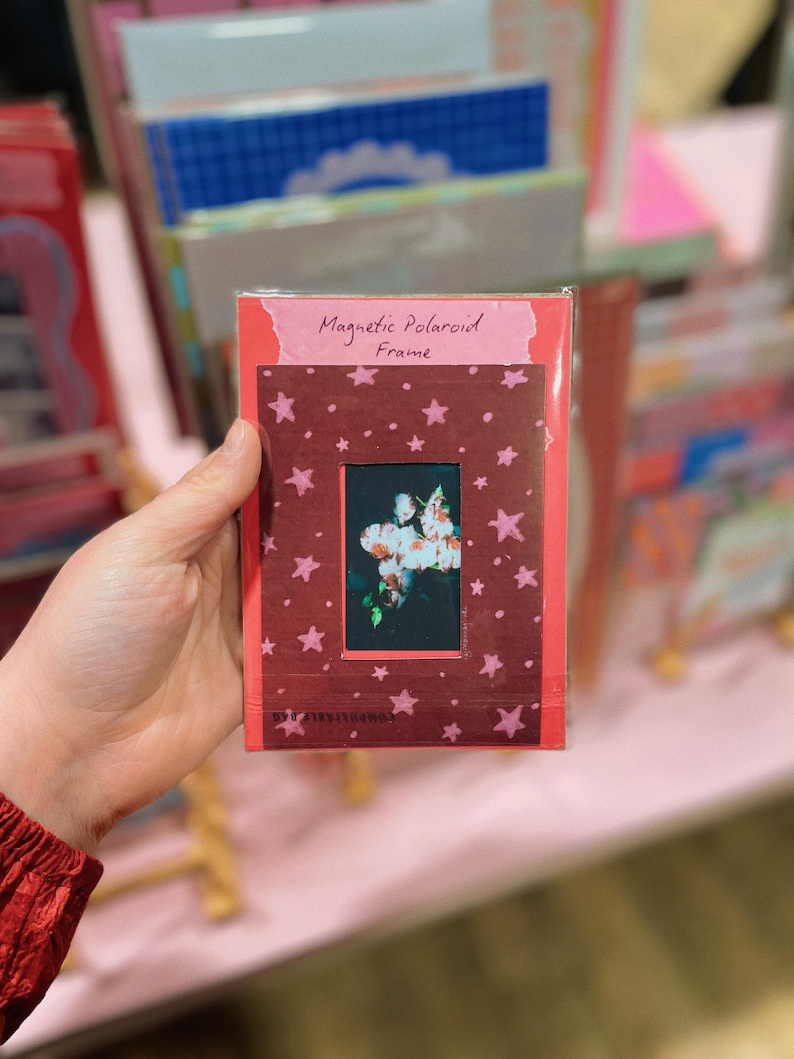 May include: A rectangular magnetic Polaroid frame in a dark red color featuring a pattern of small pink stars. The frame has a central rectangular cutout displaying a dark image of white flowers. The text Magnetic Polaroid Frame is printed at the top.