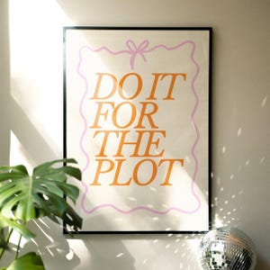 May include: A framed art print featuring the phrase "DO IT FOR THE PLOT" in orange on a cream background. The text is enclosed by a wavy pink border and a bow. The print is presented in a black frame.