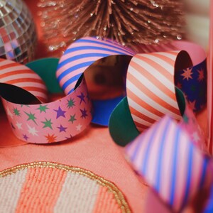 May include: A close-up of colourful paper ribbons with striped and star patterns. The ribbons are in shades of pink, blue, and orange, creating a cheerful vibe. A disco ball and a decorative tree are in the background.