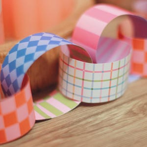 May include: A decorative paper chain featuring a variety of patterns, such as pink stripes, blue diamond shapes, and orange and pink chequered designs. The chain is arranged in a circular form, with the patterned paper creating a visual effect. The background is a blurred wooden surface.