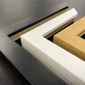May include: Three picture frames, one black, one white, and one wood, are stacked on top of each other. The frames are all different sizes and have different styles.