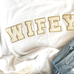 May include: White sweatshirt with the word "WIFEY" in gold and white felt letters.
