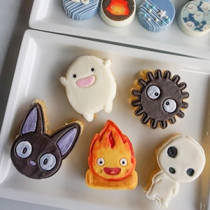 May include: A collection of decorated cookies and treats, arranged on white rectangular platters. The cookies feature various designs, including cartoon characters, blue frosting, and white sprinkles. Pink flowers are in the background.