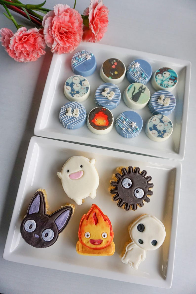 May include: A collection of decorated cookies and treats, arranged on white rectangular platters. The cookies feature various designs, including cartoon characters, blue frosting, and white sprinkles. Pink flowers are in the background.