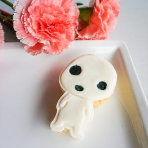 May include: A white cookie shaped like a character with black eyes and a round head, placed on a white plate. Two coral-colored carnation flowers are in the background. The cookie has a light-colored base.