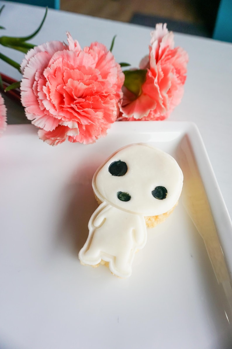 May include: A white cookie shaped like a character with black eyes and a round head, placed on a white plate. Two coral-colored carnation flowers are in the background. The cookie has a light-colored base.