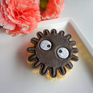 May include: A dark brown, circular cookie with a cartoon face, featuring white eyes, sits on a white plate. Pink carnations are in the background. The cookie has a textured, yellow base.
