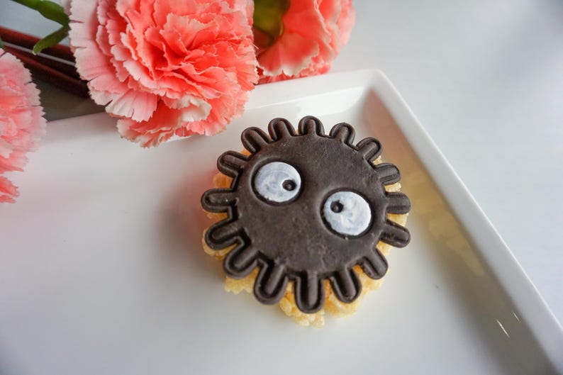 May include: A dark brown, circular cookie with a cartoon face, featuring white eyes, sits on a white plate. Pink carnations are in the background. The cookie has a textured, yellow base.
