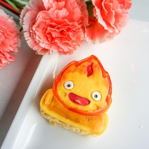 May include: A colorful, cartoon-style cookie shaped like a flame, with a smiling face and a yellow and orange color scheme. The cookie sits on a small, square base. Pink carnations are in the background.