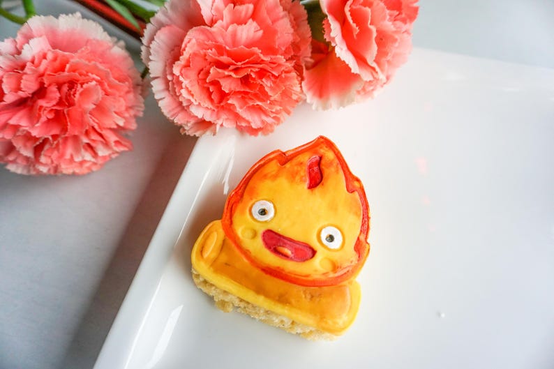 May include: A colorful, cartoon-style cookie shaped like a flame, with a smiling face and a yellow and orange color scheme. The cookie sits on a small, square base. Pink carnations are in the background.