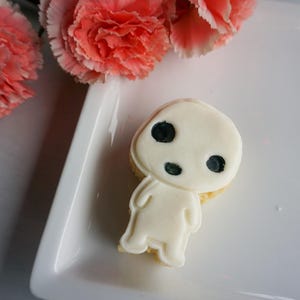 May include: A white frosted cookie shaped like a character with three black eye spots. The cookie is on a white plate, with pink carnation flowers in the background.