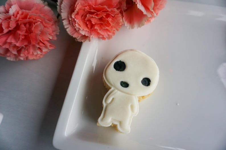 May include: A white frosted cookie shaped like a character with three black eye spots. The cookie is on a white plate, with pink carnation flowers in the background.