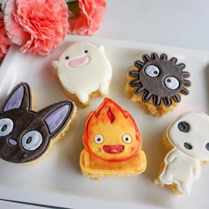May include: A collection of decorated cookies in various shapes and colors, including a black cat, a fire demon, and a soot sprite. The cookies are arranged on a white rectangular plate, with pink flowers in the background.