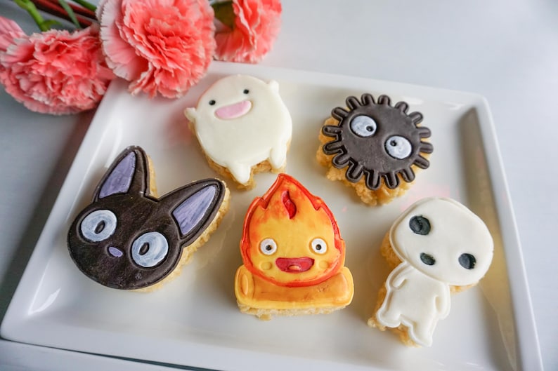 May include: A collection of decorated cookies in various shapes and colors, including a black cat, a fire demon, and a soot sprite. The cookies are arranged on a white rectangular plate, with pink flowers in the background.