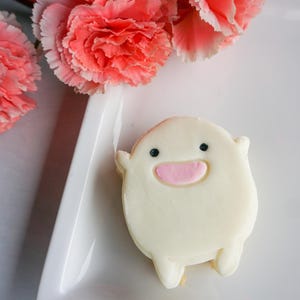 May include: A whimsical, white frosted cookie shaped like a cartoon character with black eyes and a pink mouth. The cookie rests on a white rectangular plate, with pink carnation flowers in the background.