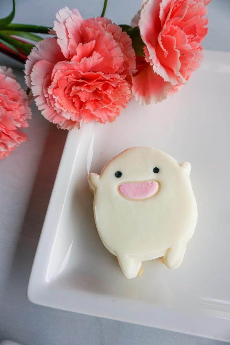 May include: A whimsical, white frosted cookie shaped like a cartoon character with black eyes and a pink mouth. The cookie rests on a white rectangular plate, with pink carnation flowers in the background.