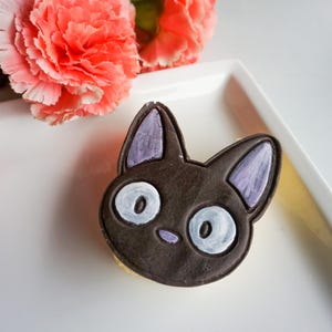 May include: A black cat-shaped cookie with white eyes and purple ears sits on a white plate, next to pink carnation flowers. The cookie is a detailed, edible treat, perfect for a themed party or a sweet gift.