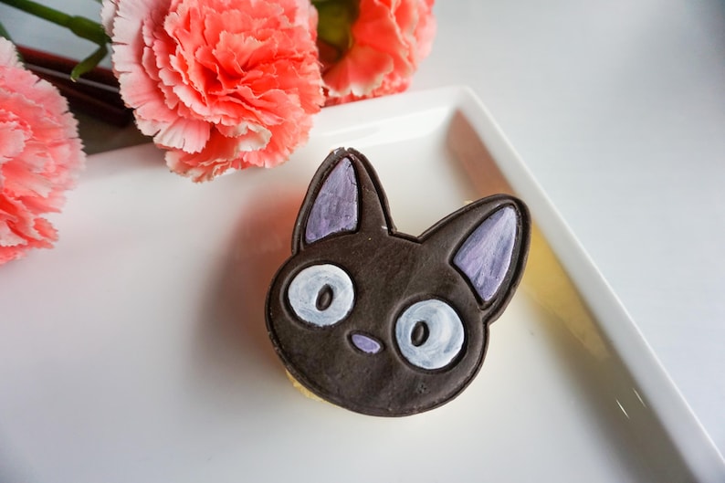 May include: A black cat-shaped cookie with white eyes and purple ears sits on a white plate, next to pink carnation flowers. The cookie is a detailed, edible treat, perfect for a themed party or a sweet gift.