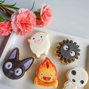 May include: A white rectangular plate holds six decorated cookies. The cookies feature cartoon characters in various shapes and colors, including a black cat, a white spirit, and a fiery creature. Pink carnations are in the background.