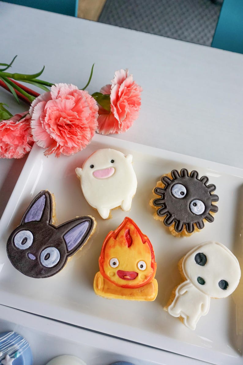 May include: A white rectangular plate holds six decorated cookies. The cookies feature cartoon characters in various shapes and colors, including a black cat, a white spirit, and a fiery creature. Pink carnations are in the background.