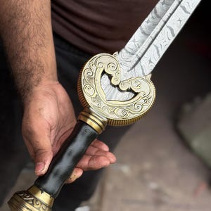 May include: A decorative sword with a patterned blade and ornate gold-colored hilt. The hilt features a black leather grip and intricate scrollwork. The sword is held in a person's hand.