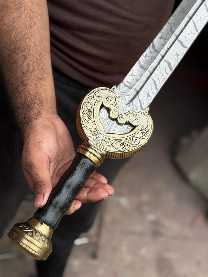 May include: A decorative sword with a patterned blade and ornate gold-colored hilt. The hilt features a black leather grip and intricate scrollwork. The sword is held in a person's hand.