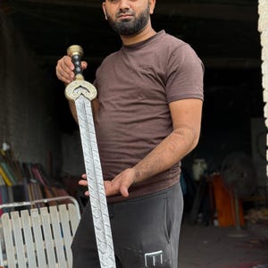 May include: A decorative sword with a detailed blade and ornate handle. The sword features a silver blade with a patterned design and a gold-colored handle with black accents. The sword is held by a person.