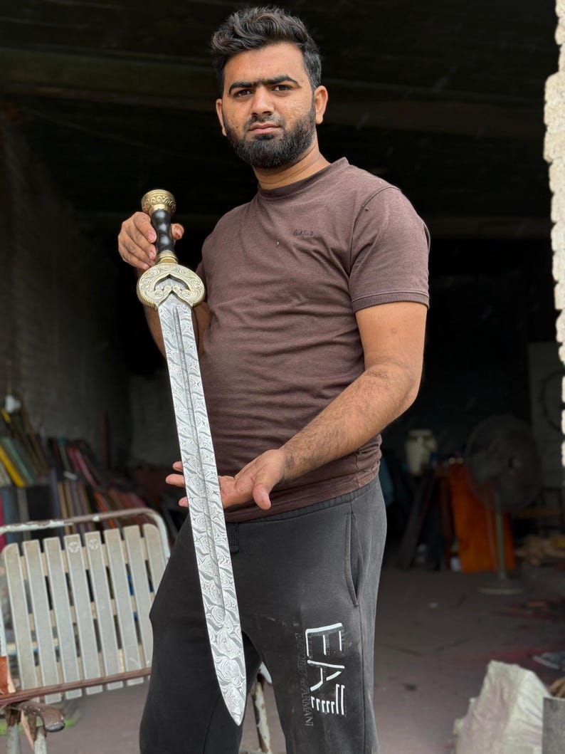 May include: A decorative sword with a detailed blade and ornate handle. The sword features a silver blade with a patterned design and a gold-colored handle with black accents. The sword is held by a person.